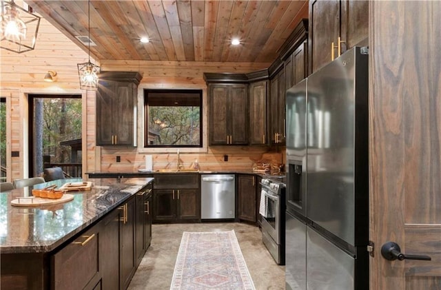 kitchen featuring appliances with stainless steel finishes, dark brown cabinets, wooden walls, pendant lighting, and dark stone countertops