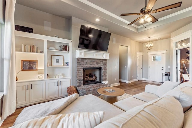 living room featuring ornamental molding, a brick fireplace, ceiling fan with notable chandelier, a raised ceiling, and light wood-type flooring