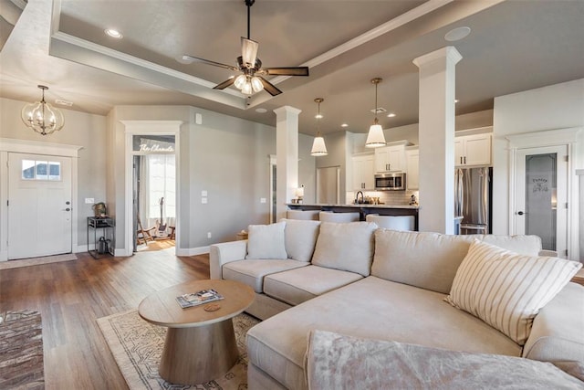 living room featuring a raised ceiling, ceiling fan with notable chandelier, decorative columns, and light wood-type flooring