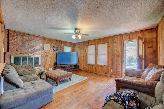 living room with wooden walls, a brick fireplace, light hardwood / wood-style flooring, ceiling fan, and a textured ceiling