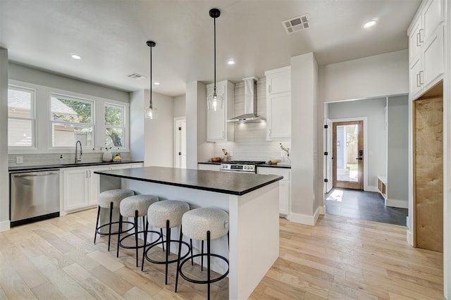 kitchen featuring a center island, hanging light fixtures, wall chimney range hood, stainless steel appliances, and white cabinets