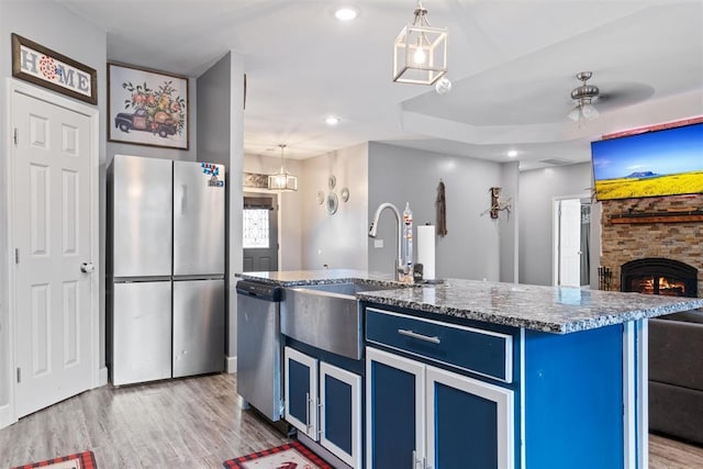 kitchen featuring a stone fireplace, blue cabinets, ceiling fan, an island with sink, and appliances with stainless steel finishes