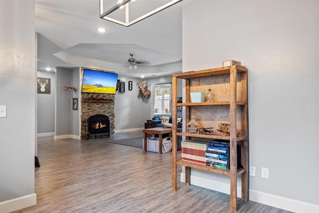 interior space with wood finished floors, baseboards, ceiling fan, a stone fireplace, and a raised ceiling