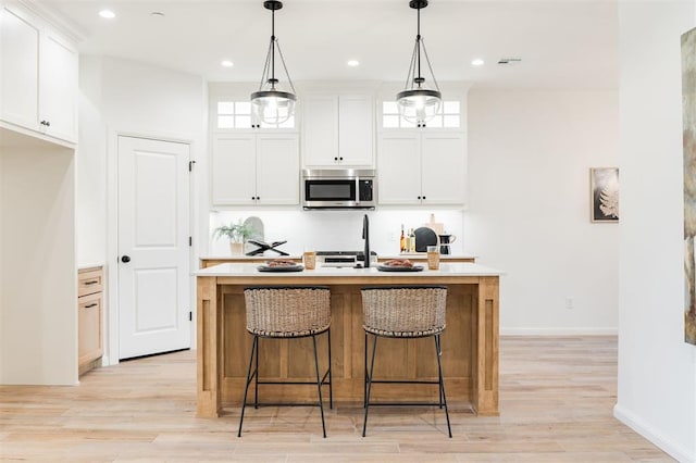 kitchen with light countertops, stainless steel microwave, light wood-style flooring, a kitchen island with sink, and white cabinets