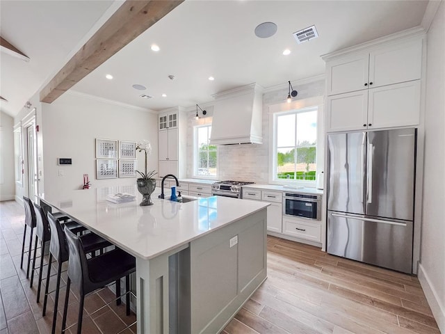 kitchen featuring stainless steel appliances, a sink, light wood-type flooring, custom exhaust hood, and a large island with sink