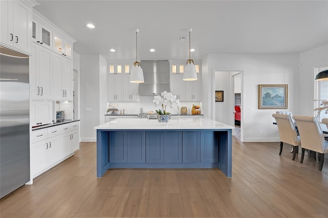 kitchen with pendant lighting, stainless steel built in refrigerator, white cabinets, wall chimney range hood, and a center island with sink