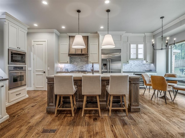 kitchen with built in appliances, a kitchen island with sink, dark wood-type flooring, and decorative light fixtures