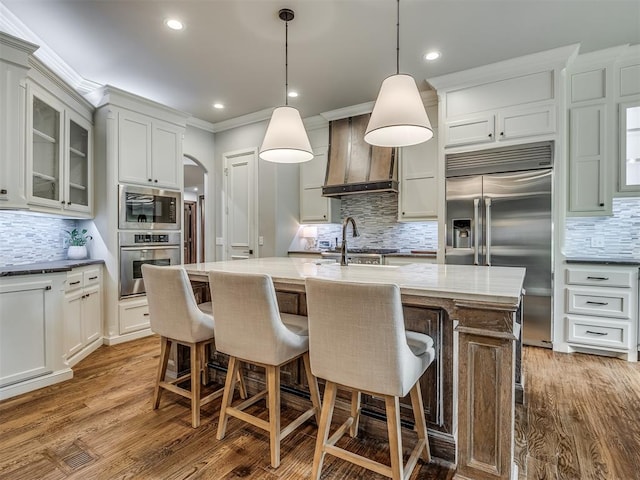 kitchen featuring built in appliances, decorative light fixtures, an island with sink, and tasteful backsplash