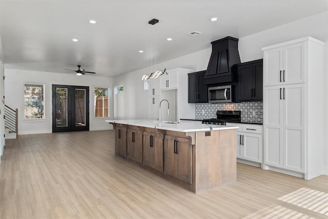 kitchen featuring a center island with sink, black stove, hanging light fixtures, light wood-type flooring, and white cabinetry