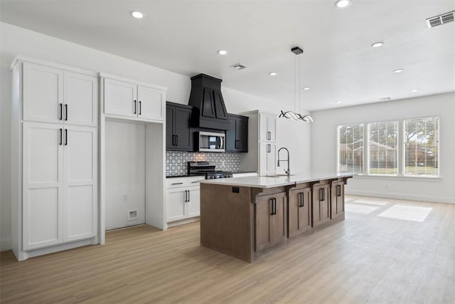 kitchen with pendant lighting, a kitchen island with sink, white cabinets, light wood-type flooring, and appliances with stainless steel finishes