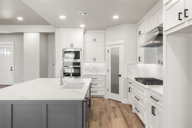 kitchen with wall chimney exhaust hood, light stone countertops, white cabinets, and an island with sink