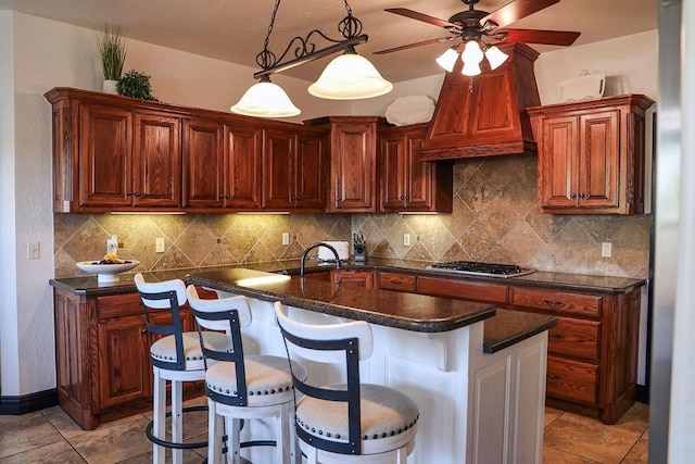 kitchen with decorative backsplash, stainless steel gas cooktop, ceiling fan, dark countertops, and a kitchen island with sink