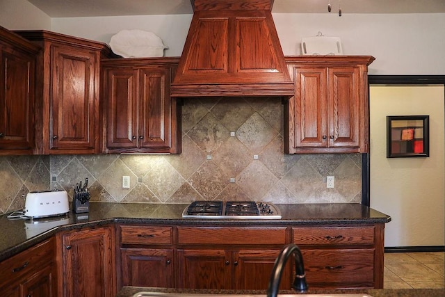 kitchen featuring stainless steel gas stovetop, decorative backsplash, light tile patterned flooring, custom exhaust hood, and dark stone counters