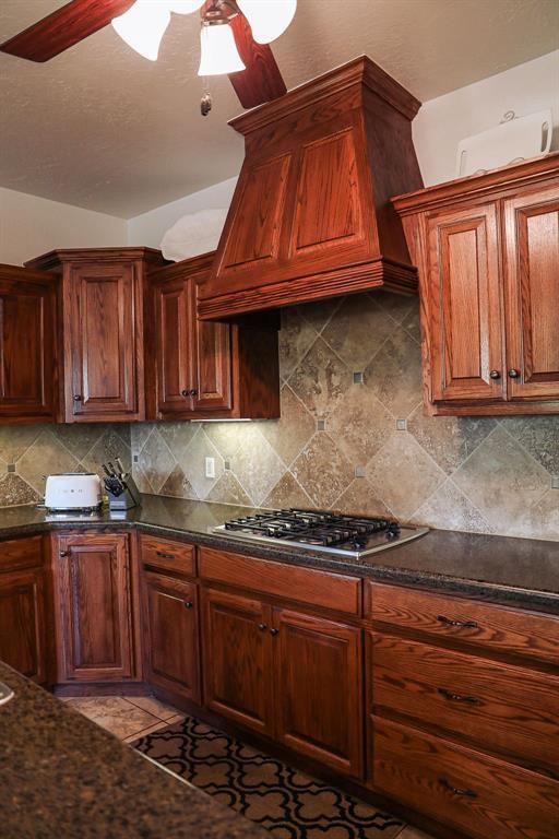 kitchen with custom exhaust hood, stainless steel gas cooktop, ceiling fan, tasteful backsplash, and dark stone countertops