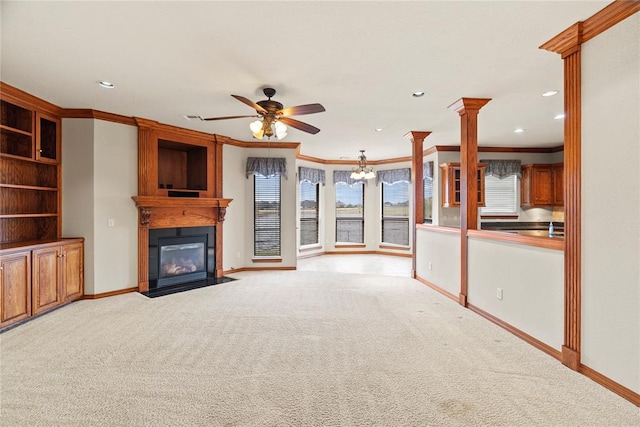 unfurnished living room featuring ceiling fan with notable chandelier, crown molding, a large fireplace, light colored carpet, and decorative columns