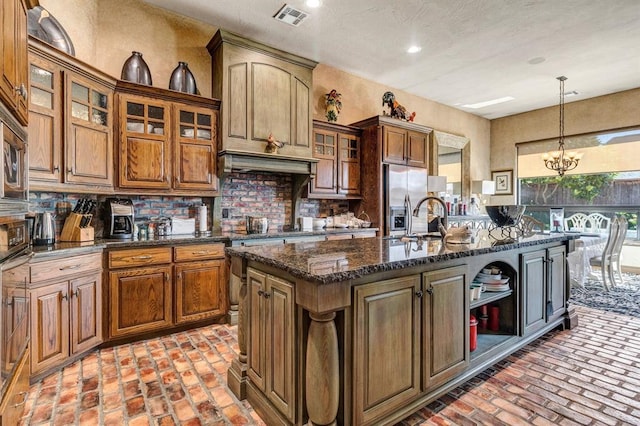 kitchen featuring a kitchen island with sink, an inviting chandelier, stainless steel refrigerator with ice dispenser, dark stone countertops, and decorative light fixtures