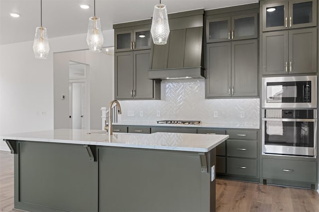 729 Caladium Dr, Yukon, OK 73099 - kitchen featuring backsplash, custom range hood, wood finished floors, stainless steel appliances, and a kitchen island with sink