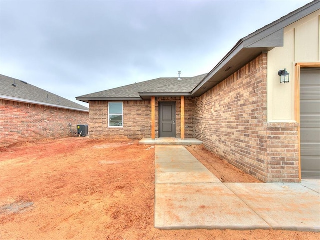 view of exterior entry with an attached garage, a shingled roof, central AC, and brick siding