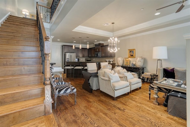 living room with ceiling fan with notable chandelier, a raised ceiling, light wood-type flooring, and crown molding