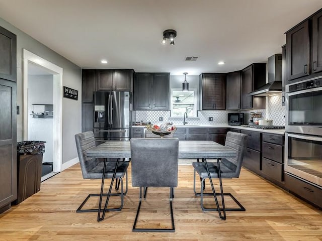 kitchen featuring backsplash, wall chimney range hood, light wood-type flooring, dark brown cabinetry, and stainless steel appliances