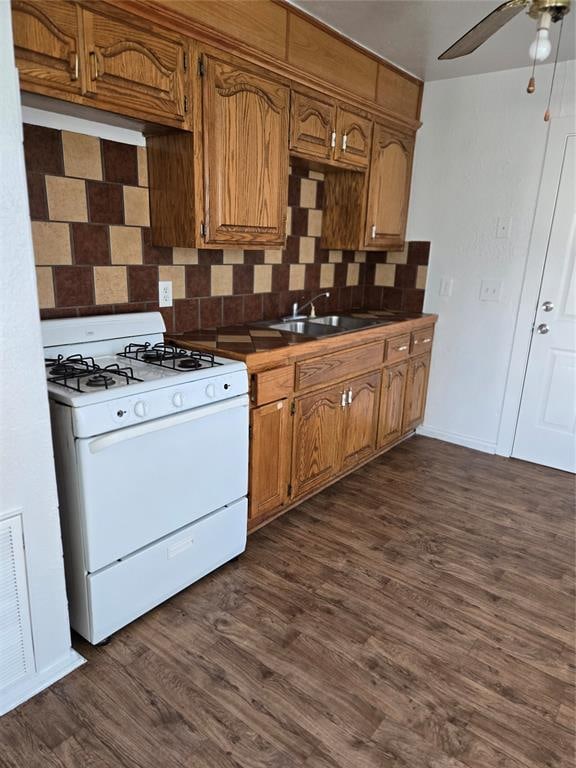 kitchen featuring white gas range oven, brown cabinetry, decorative backsplash, dark wood-type flooring, and a sink