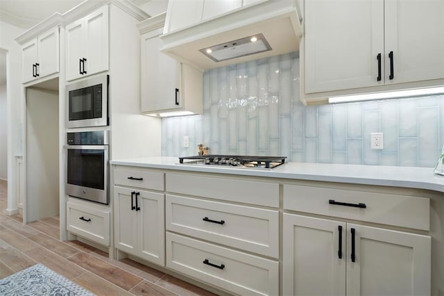 kitchen with premium range hood, light hardwood / wood-style floors, decorative backsplash, white cabinets, and appliances with stainless steel finishes