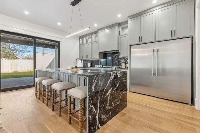 kitchen with an island with sink, dark stone counters, pendant lighting, stainless steel built in fridge, and light wood-type flooring