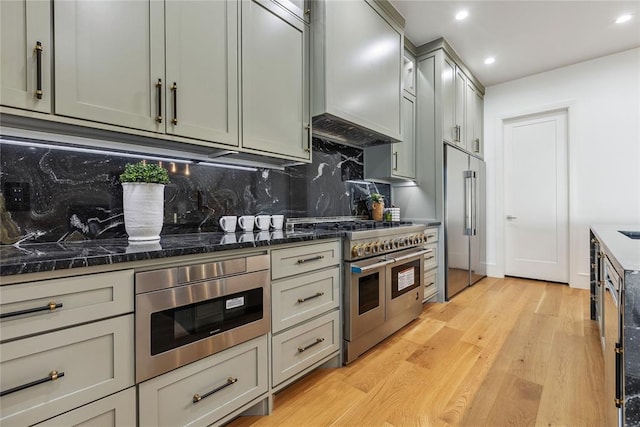 kitchen featuring tasteful backsplash, premium range hood, high end appliances, light hardwood / wood-style flooring, and dark stone countertops