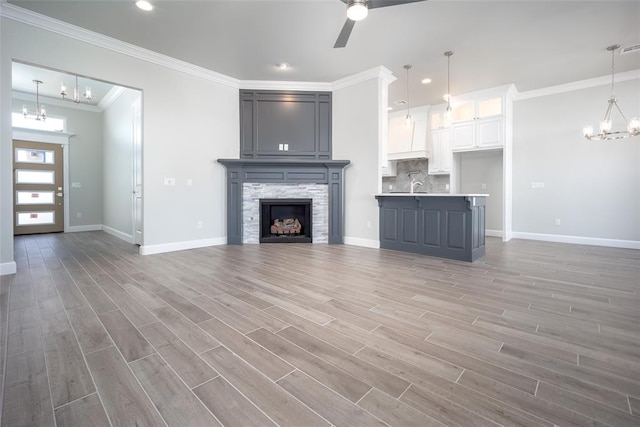 unfurnished living room with ornamental molding, ceiling fan with notable chandelier, sink, light hardwood / wood-style floors, and a stone fireplace