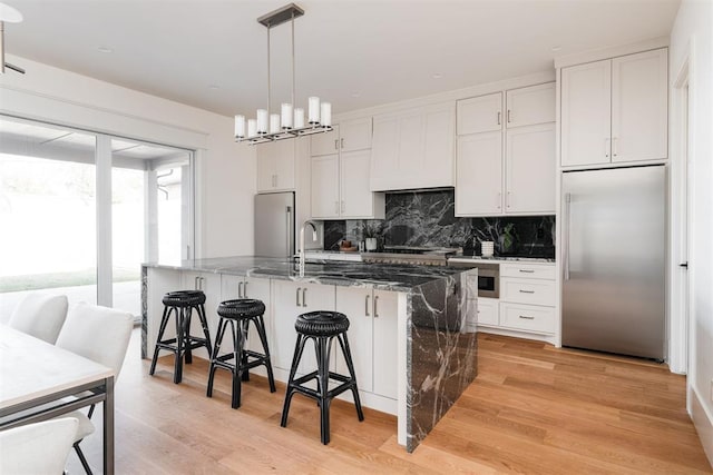 kitchen featuring white cabinets, stainless steel built in fridge, a kitchen island with sink, and dark stone counters