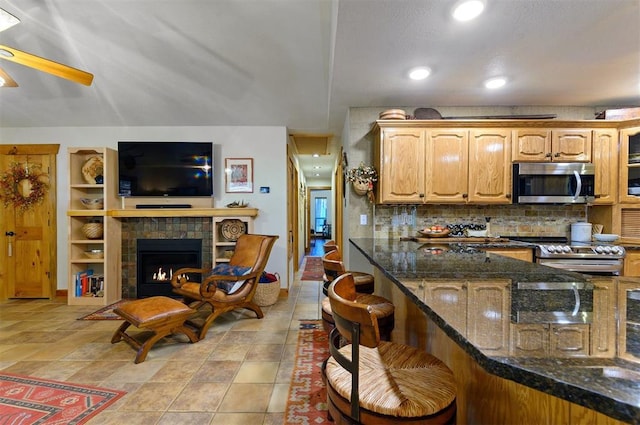 kitchen with ceiling fan, dark stone counters, a tiled fireplace, a breakfast bar, and appliances with stainless steel finishes