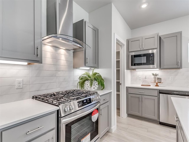 kitchen with wall chimney range hood, light hardwood / wood-style flooring, gray cabinets, appliances with stainless steel finishes, and tasteful backsplash