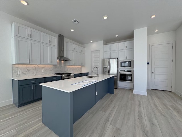 kitchen featuring sink, light wood-type flooring, a kitchen island with sink, stainless steel appliances, and wall chimney exhaust hood