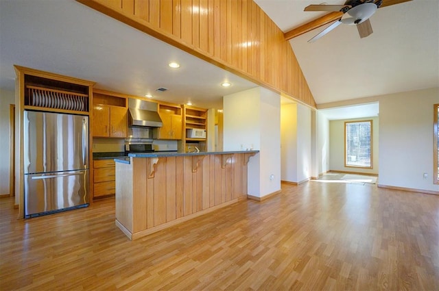 kitchen with wall chimney range hood, ceiling fan, light wood-type flooring, appliances with stainless steel finishes, and a breakfast bar area