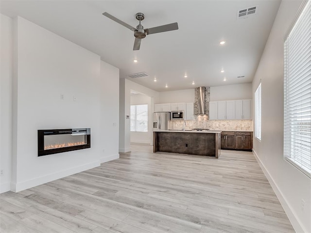 kitchen featuring appliances with stainless steel finishes, wall chimney range hood, white cabinets, an island with sink, and backsplash