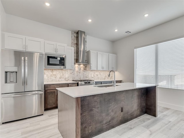 kitchen featuring appliances with stainless steel finishes, wall chimney exhaust hood, sink, light wood-type flooring, and a kitchen island with sink