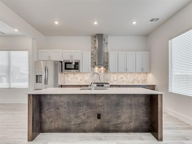 kitchen featuring built in microwave, a kitchen island with sink, stainless steel refrigerator with ice dispenser, wall chimney range hood, and sink