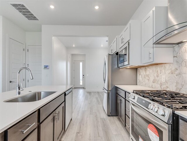 kitchen featuring appliances with stainless steel finishes, sink, white cabinetry, wall chimney range hood, and light hardwood / wood-style floors