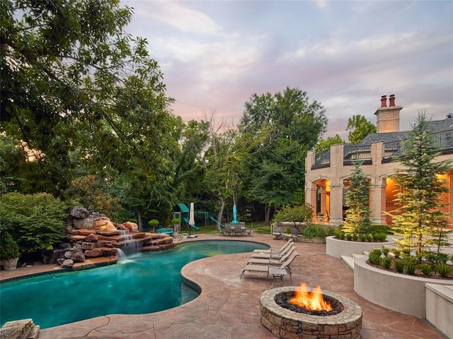 pool at dusk featuring pool water feature, a patio, and an outdoor fire pit