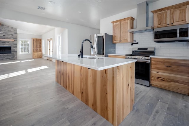 kitchen featuring backsplash, a kitchen island with sink, wall chimney range hood, light hardwood / wood-style flooring, and appliances with stainless steel finishes