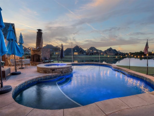 pool at dusk with a patio area, an in ground hot tub, a water view, and an outdoor fireplace