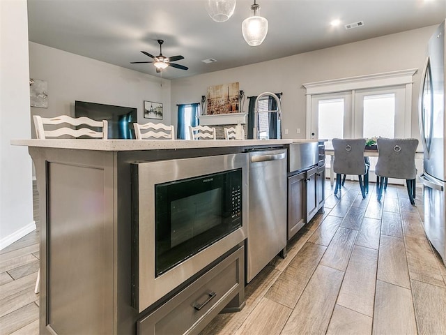 kitchen with ceiling fan, stainless steel appliances, pendant lighting, a center island with sink, and light wood-type flooring