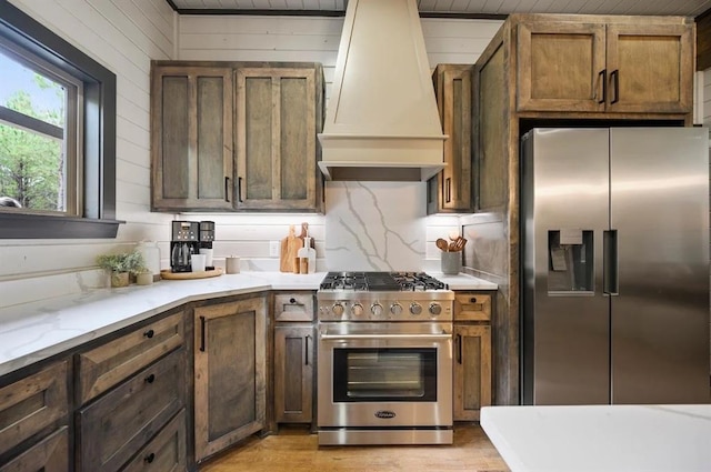 kitchen featuring stainless steel appliances, tasteful backsplash, light stone counters, custom range hood, and light wood-type flooring