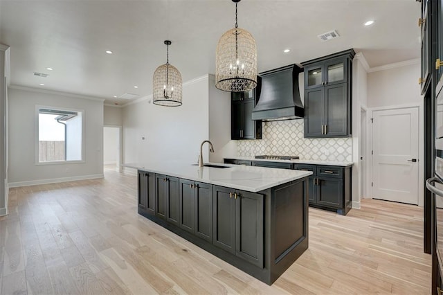 kitchen featuring light wood-type flooring, custom exhaust hood, crown molding, pendant lighting, and a center island with sink