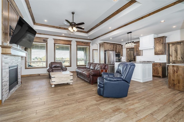 living room featuring ceiling fan, a stone fireplace, light hardwood / wood-style floors, a tray ceiling, and ornamental molding