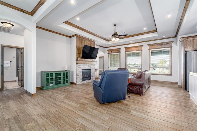 living room with ornamental molding, a raised ceiling, ceiling fan, light hardwood / wood-style floors, and a stone fireplace