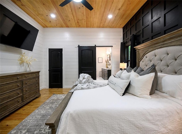 bedroom with ensuite bath, ceiling fan, a barn door, light hardwood / wood-style flooring, and wood walls