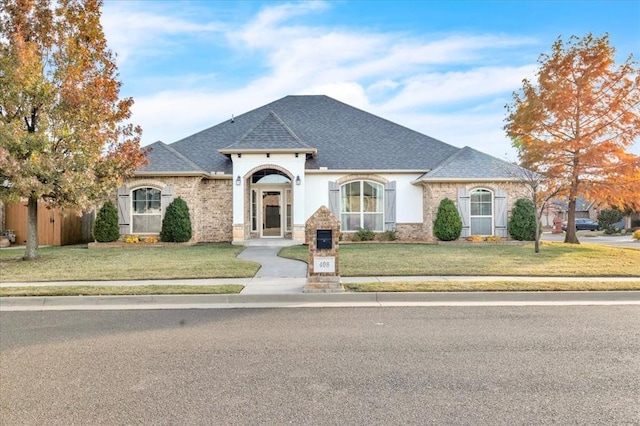 french country inspired facade featuring a front yard, roof with shingles, fence, and brick siding