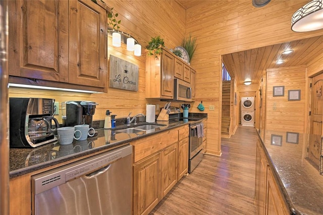 kitchen with stainless steel appliances, sink, light hardwood / wood-style flooring, stacked washer / drying machine, and wood walls