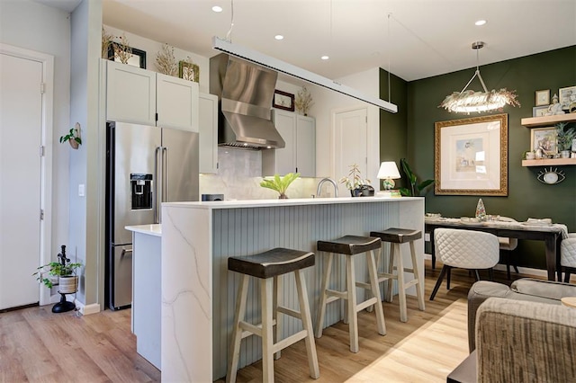 kitchen with high end refrigerator, wall chimney range hood, decorative light fixtures, light hardwood / wood-style floors, and white cabinetry